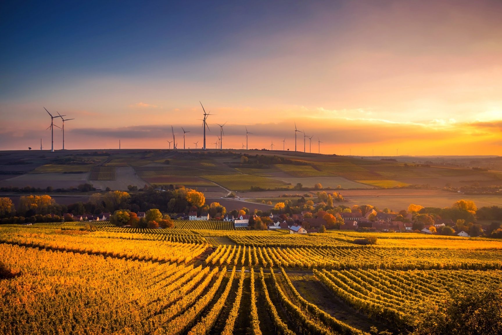 A landscape at sunset. There is a field of crops, a small valley with a village, and wind turbines in the distance.