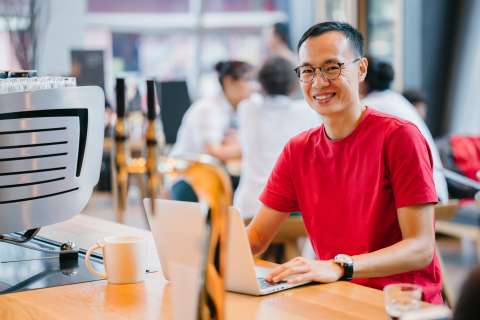 A man wearing glasses in a red t-shirt at a coffee shop working at a laptop. He is smiling at the camera.