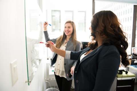 Two women at work. They are working together at a whiteboard. They are smiling.